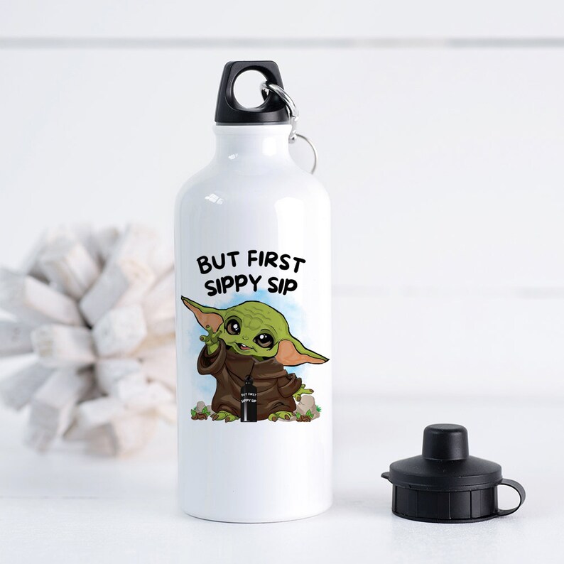 May include: White aluminum water bottle with a black lid and a black carrying loop. The bottle has a graphic of a green alien character holding a smaller bottle with the text "But First Sippy Sip".