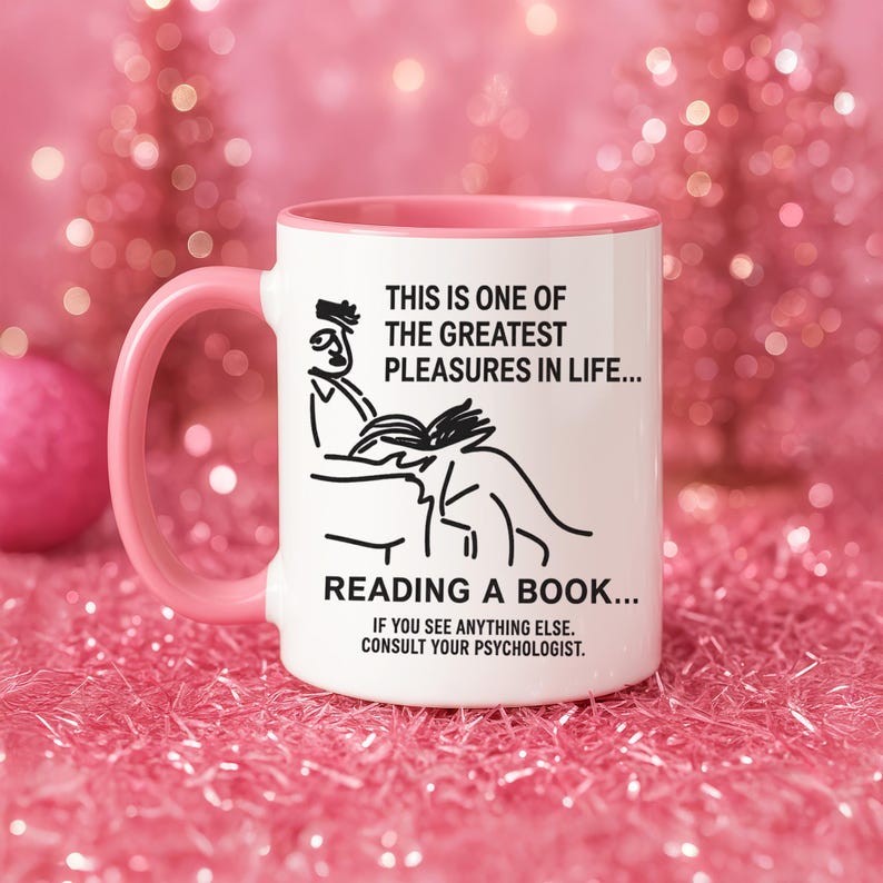 May include: White ceramic mug with a pink interior and handle. The mug features a black line drawing of a person reading a book, with the text "THIS IS ONE OF THE GREATEST PLEASURES IN LIFE... READING A BOOK..." and "IF YOU SEE ANYTHING ELSE. CONSULT YOUR PSYCHOLOGIST."