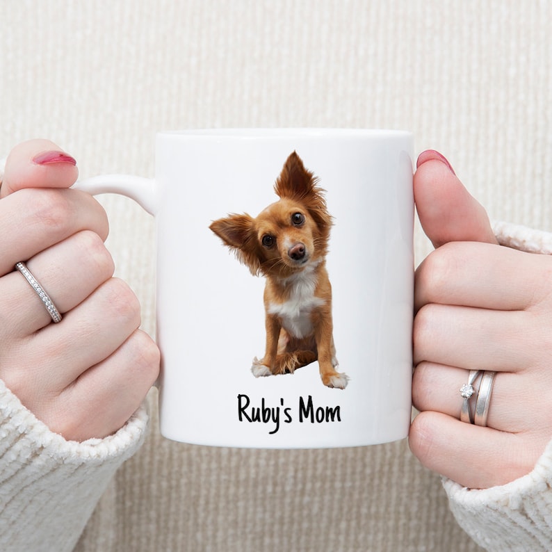 May include: White ceramic mug featuring a brown and white dog. The dog is looking at the camera with its head tilted. The text "Ruby's Mom" is printed on the mug.