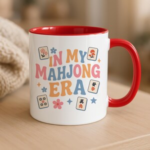 May include: White ceramic mug with a red interior and handle. The mug features the text "IN MY MAHONG ERA" in colorful, retro-style lettering, along with Mahjong tile illustrations and floral accents. A fun and stylish mug for game lovers.
