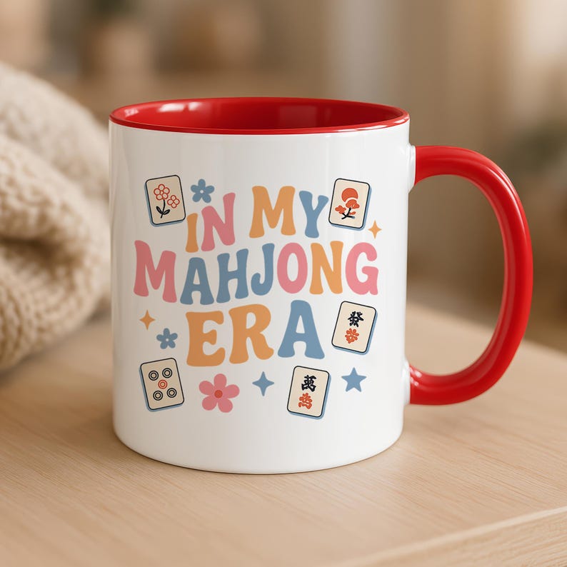 May include: White ceramic mug with a red interior and handle. The mug features the text "IN MY MAHONG ERA" in colorful, retro-style lettering, along with Mahjong tile illustrations and floral accents. A fun and stylish mug for game lovers.