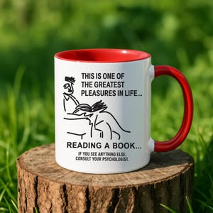 May include: White ceramic mug with a red interior and handle. The mug features a black illustration of a person reading a book, with text that reads "THIS IS ONE OF THE GREATEST PLEASURES IN LIFE... READING A BOOK... IF YOU SEE ANYTHING ELSE. CONSULT YOUR PSYCHOLOGIST."