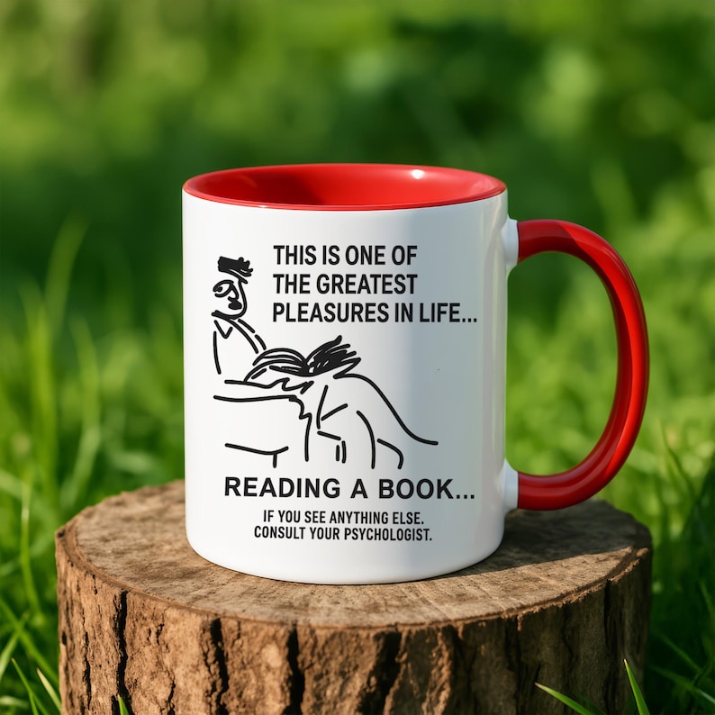 May include: White ceramic mug with a red interior and handle. The mug features a black illustration of a person reading a book, with text that reads "THIS IS ONE OF THE GREATEST PLEASURES IN LIFE... READING A BOOK... IF YOU SEE ANYTHING ELSE. CONSULT YOUR PSYCHOLOGIST."