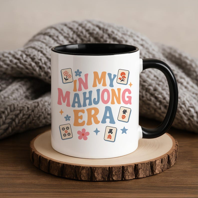 May include: White ceramic mug with a black interior and handle. The mug features the text "IN MY MAHONG ERA" in a retro font, along with floral and tile graphics. The mug sits on a wooden coaster.