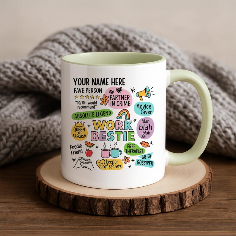 May include: A white ceramic mug with a light green handle and interior. The mug features colorful text and illustrations, including "Work Bestie," "Fave Person," and other fun phrases. The mug sits on a wooden coaster.