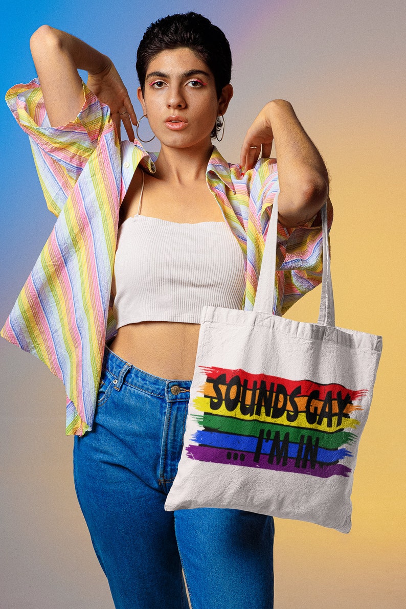 May include: A person wearing a striped shirt, a white vest top, and blue jeans is holding a white tote bag with the text "Sounds Gay I'm In" printed on it in black. The text is surrounded by a rainbow stripe.