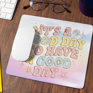 Peut inclure: Un tapis de souris rose et bleu avec un fond dégradé arc-en-ciel. Le texte "It's a good day to have a good day" est écrit dans une police rétro avec des fleurs et des étoiles.