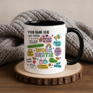 May include: White ceramic mug with a black interior and handle. The mug features colorful text and graphics, including "Work Bestie," "Partner in Crime," and "Free Therapist." The mug sits on a wooden coaster.