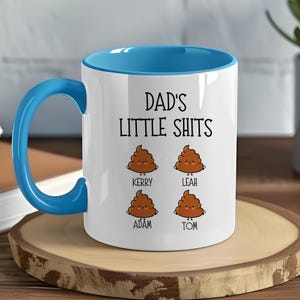 Op de afbeelding: Witte keramische mok met een blauwe binnenkant en handvat. De mok heeft de tekst "DAD'S LITTLE SHITS" en vier cartoon poep-emoji's met namen: Kerry, Leah, Adam en Tom. De mok staat op een houten onderzetter.