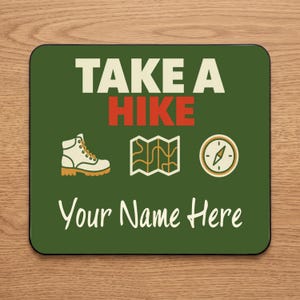 May include: A rectangular mouse pad with a dark green background featuring the text TAKE A HIKE in white and red. Below the text are icons of a hiking boot, a folded map, and a compass. The bottom of the mouse pad displays the text Your Name Here.