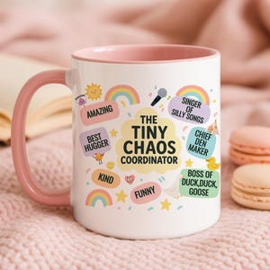 Może przedstawiać: Biały ceramiczny kubek z różowym uchwytem i wnętrzem. Kubek zawiera kolorowy tekst i ilustracje, w tym słowa "The Tiny Chaos Coordinator", tęcze i gwiazdy. Inne frazy to "Amazing", "Best Hugger" i "Funny".