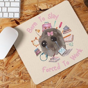 May include: A light pink and cream mouse mat with a cartoon illustration of a grey creature surrounded by school supplies. The text "Born to Slay Forced to Work" is written in pink letters.