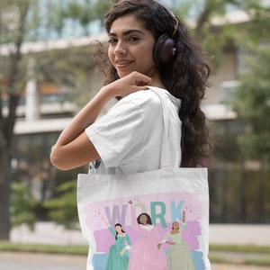 Op de afbeelding: Een witte tote bag met een roze, blauwe en groene illustratie van drie vrouwen in jurken. De tekst "WORK" is in regenboogkleuren op de tas gedrukt.