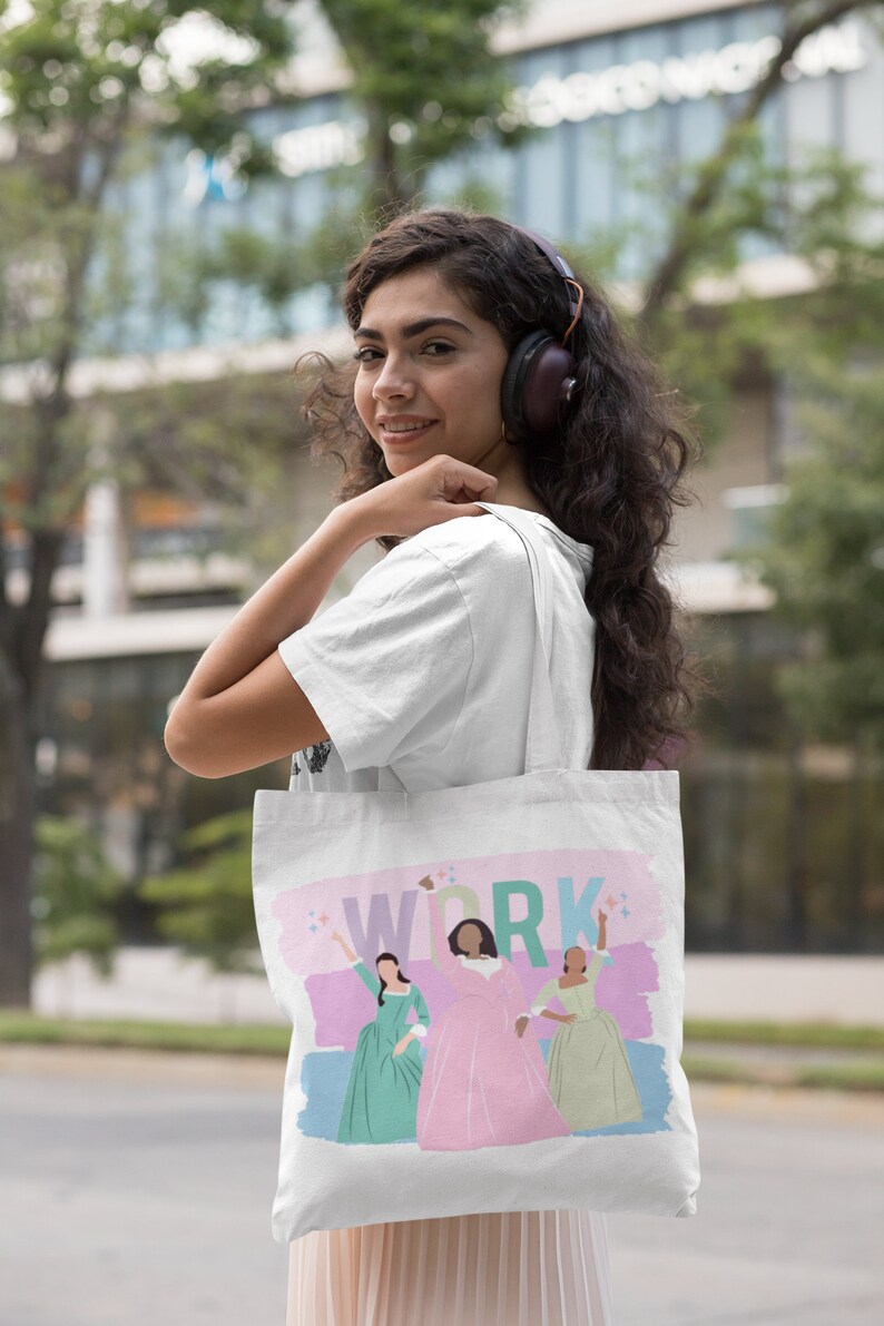 Op de afbeelding: Een witte tote bag met een roze, blauwe en groene illustratie van drie vrouwen in jurken. De tekst "WORK" is in regenboogkleuren op de tas gedrukt.