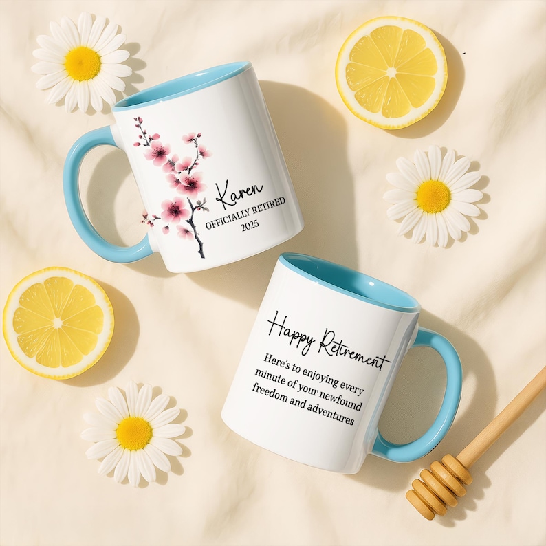 May include: Two white ceramic mugs with light blue handles and rims. One mug has a floral design and the name "Karen" with "OFFICIALLY RETIRED 2025". The other mug says "Happy Retirement". Daisies and lemon slices are scattered around.