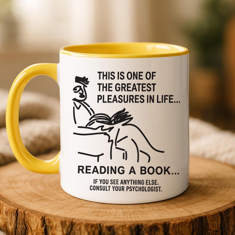 May include: White ceramic mug with a yellow interior and handle. The mug features black line art of a person reading and text that reads, "THIS IS ONE OF THE GREATEST PLEASURES IN LIFE... READING A BOOK... IF YOU SEE ANYTHING ELSE. CONSULT YOUR PSYCHOLOGIST."