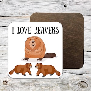 Könnte beinhalten: Ein weißer Untersetzer mit dem Text "I LOVE BEAVERS" in Schwarz. Der Untersetzer zeigt einen Cartoon-Biber mit braunem Körper und weißem Bauch. Zwei kleinere Biber befinden sich unter dem größeren Biber. Der Untersetzer hat eine braune Korkrückseite.