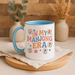 May include: White ceramic mug with a light blue interior and handle. The mug features the text "IN MY MAHJONG ERA" in colorful retro lettering, along with Mahjong tile illustrations and floral accents. The mug sits on a wooden board.