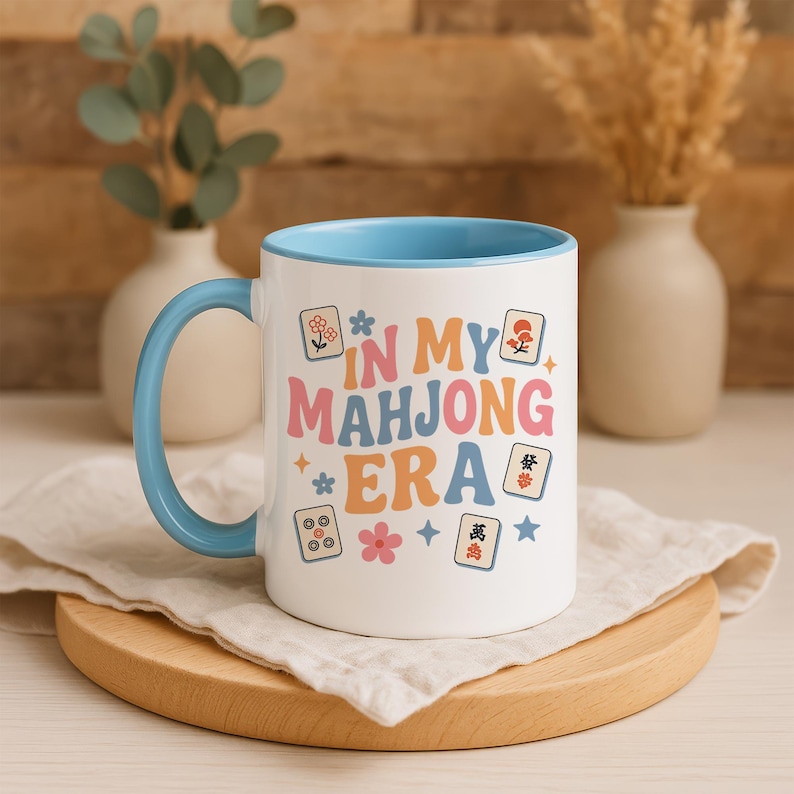 May include: White ceramic mug with a light blue interior and handle. The mug features the text "IN MY MAHJONG ERA" in colorful retro lettering, along with Mahjong tile illustrations and floral accents. The mug sits on a wooden board.