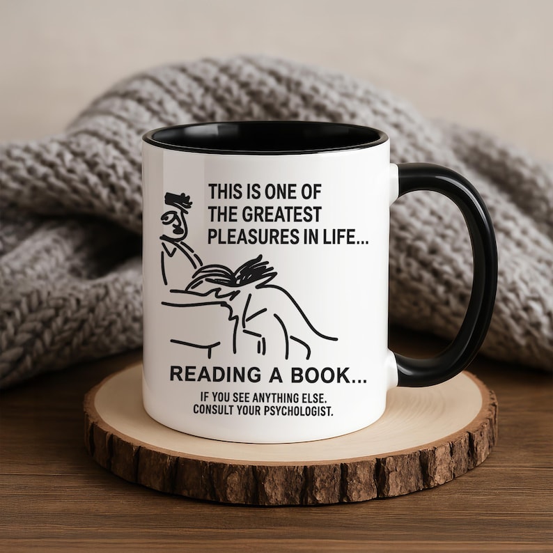 May include: White ceramic mug with a black handle and interior. The mug features a black and white illustration of a person reading a book, with the text "THIS IS ONE OF THE GREATEST PLEASURES IN LIFE... READING A BOOK..." and a humorous message.