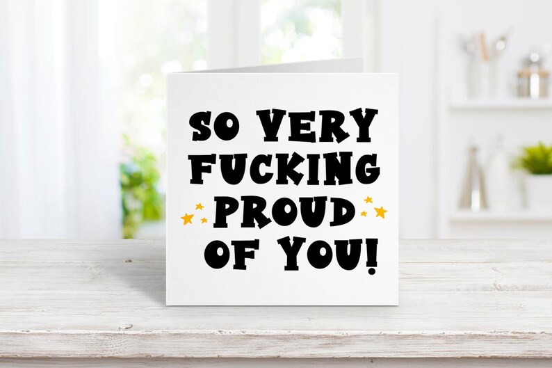 以下が含まれることがあります： 「So very fucking proud of you!」という黒い文字が書かれた白いカード。proudの両側に金色の星が2つある。