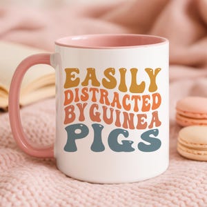May include: White ceramic mug with a pink handle and interior. The mug features the text "EASILY DISTRACTED BY GUINEA PIGS" in orange, yellow, and blue lettering. The mug is sitting on a pink surface.