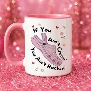 May include: White ceramic mug with a pink handle and interior. The mug features the text "If You Ain't Crocin' You Ain't Rockin'" with a graphic of a pink clog shoe. Decorative hearts, flowers, and stars are also included.