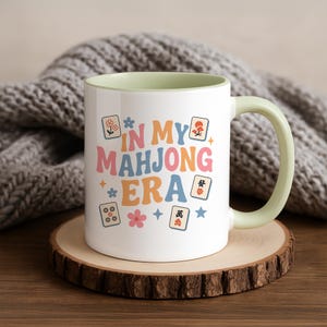 May include: White ceramic mug with a light green interior and handle. The mug features the text "IN MY MAHJONG ERA" in pastel colors, along with Mahjong tile illustrations and floral accents. The mug sits on a wooden coaster.