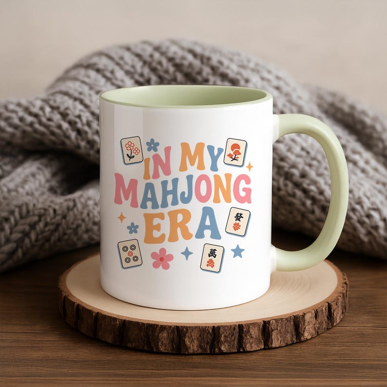 May include: White ceramic mug with a light green interior and handle. The mug features the text "IN MY MAHJONG ERA" in pastel colors, along with Mahjong tile illustrations and floral accents. The mug sits on a wooden coaster.