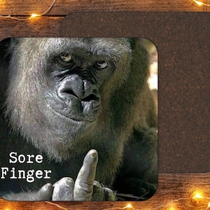 May include: Square coaster featuring a gorilla giving the middle finger. The text "Sore Finger" is printed in white. The coaster has a dark brown cork backing.