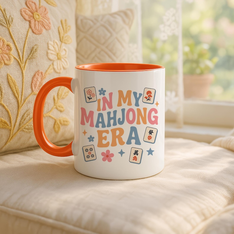 May include: White ceramic mug with an orange handle and interior. The mug features the text "IN MY MAHJONG ERA" in colorful retro lettering, along with various Mahjong tile illustrations. The mug is set against a soft, neutral background.