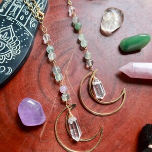 May include: A pair of gold-toned crescent moon earrings featuring dangling clear quartz points and strands of small, round, pale green and translucent beads. The earrings are constructed with thin gold metal chains and wire-wrapped crystal accents.