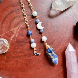 Puede incluir: Un collar de cadena en tono dorado con cuentas azules y blancas, un dije de media luna blanca, un dije pequeño de media luna azul oscuro y un colgante de piedra azul y blanca facetada envuelto en alambre dorado en la parte inferior.