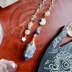 May include: A gold chain necklace with a blue and white gemstone pendant, moon and star charms, and yellow and blue beads. The necklace is displayed on a wooden surface with crystals and a decorative hand.