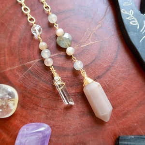May include: Close-up of two gold-colored chain necklaces with gemstone pendants. One pendant is a clear crystal, and the other is a pinkish-beige crystal. The necklaces are adorned with small, round, and faceted beads in shades of white, beige, and green. The background is a reddish-brown wooden surface.