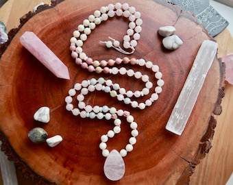 May include: A pink, white, and light blue crystal mala necklace with a large pink crystal pendant. The necklace is laid out on a wooden surface with other crystals.