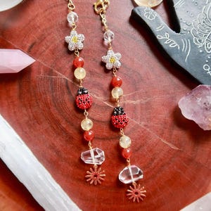 Puede incluir: Pendientes dorados con abalorios de flores y mariquitas, con cuentas rojas, amarillas y transparentes. Los pendientes se exhiben sobre una superficie de madera con cristales y un objeto decorativo en forma de mano.