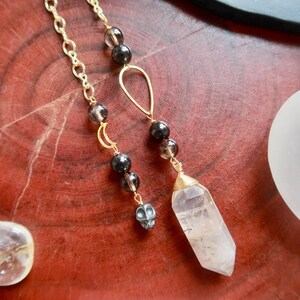 May include: Gold-colored chain earrings with smoky quartz beads and a clear quartz crystal pendant. One earring features a small skull charm. The earrings are displayed on a reddish-brown wooden surface, with crystals in the background.