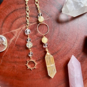 Pode incluir: Um pêndulo de corrente de metal dourado com uma ponta de cristal amarelo facetado, contas esféricas transparentes e amarelas, um anel dourado fino e um pequeno pingente em forma de sol, suspenso por delicados elos de metal dourado.