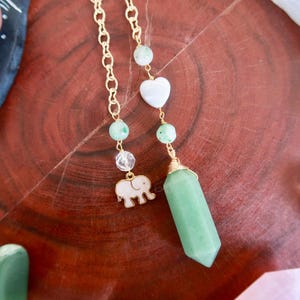 May include: A green crystal pendulum with a gold chain and beads. The pendulum is shaped like a point and has a small elephant charm attached to the chain.