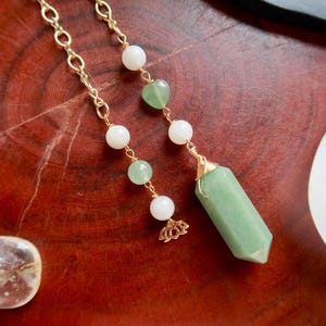 May include: Two gold-colored chain pendulums with white and green beads, and a green crystal pendant. The pendulums are on a reddish-brown surface, with clear quartz crystals and a white sphere in the background. The crystal pendant is approximately 1.5 inches long.