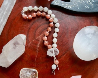 May include: A collection of crystals and a beaded mala necklace on a reddish-brown wooden surface. The necklace features white and peach-colored beads with a clear crystal pendant. Other crystals include a large clear quartz and a selenite wand.