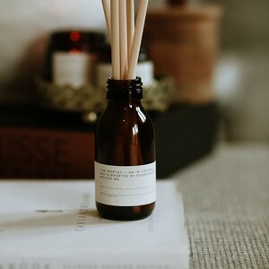 May include: A brown glass bottle with a white label that reads "I am worthy. I am in control and supported by everything around me." The bottle has wooden sticks inserted into it. The bottle is sitting on a white book.