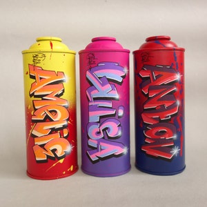 May include: Three spray paint cans with graffiti art. The cans are yellow, purple, and blue with the words "SYDNEY", "ASTIK", and "AMERICA" written in colorful graffiti style.