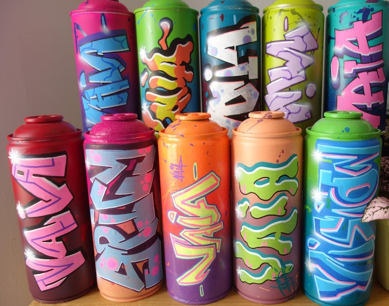 May include: A collection of ten spray paint cans with colorful graffiti designs. The cans are arranged in two rows, with five cans in each row. The cans are all different colors, including pink, red, orange, yellow, green, blue, and purple. The graffiti designs are all unique and feature a variety of letters, numbers, and symbols.