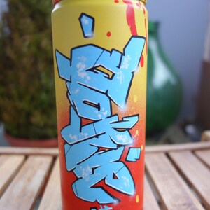 May include: A yellow, red, and blue spray paint can with graffiti-style lettering. The lettering is in a blue and white color scheme and appears to be Japanese characters.
