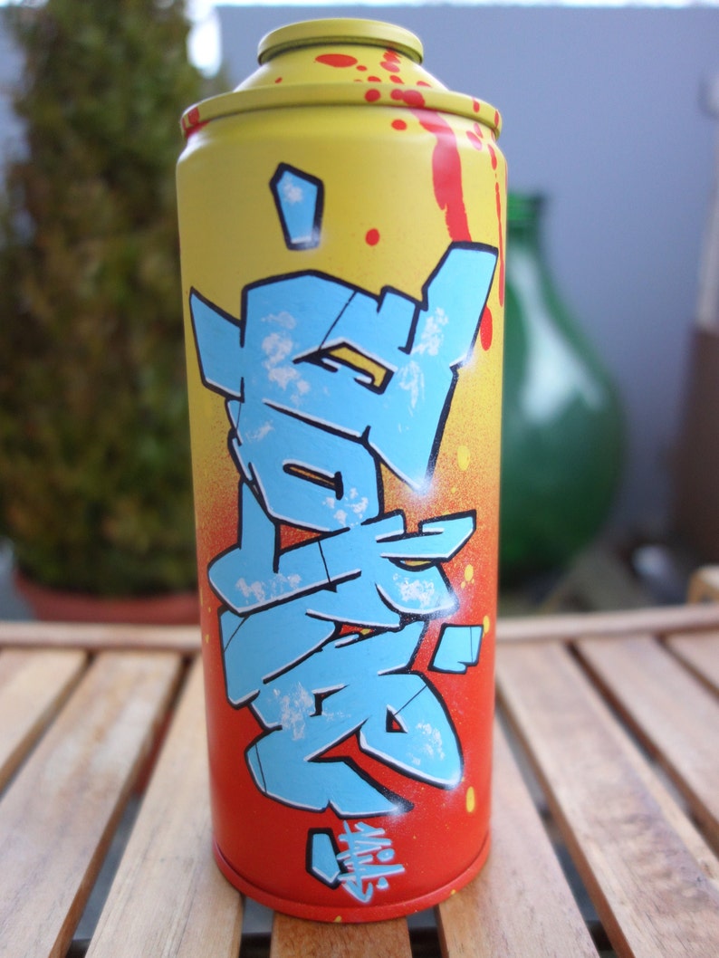 May include: A yellow, red, and blue spray paint can with graffiti-style lettering. The lettering is in a blue and white color scheme and appears to be Japanese characters.