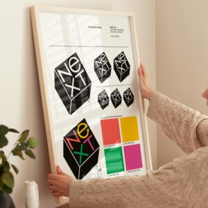 May include: A framed print featuring the word "NEXT" in a geometric design. The design is repeated in various sizes and colors, with a color palette displayed below. The print is held by a person.