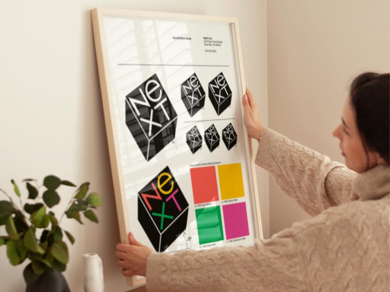 May include: A framed print featuring the word "NEXT" in a geometric design. The design is repeated in various sizes and colors, with a color palette displayed below. The print is held by a person.