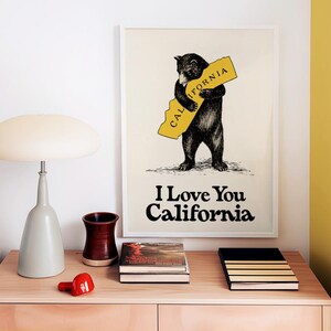 May include: A framed print with a black bear hugging a yellow outline of the state of California. The text "I Love You California" is printed below the bear. The print is on a wooden table with books and a lamp.